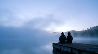 Two people sitting on wooden dock overlooking misty lake at dawn with mountains and forest in background creating peaceful morning atmosphere.