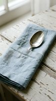 Silver spoon resting on folded blue linen napkin atop whitewashed wooden table with natural window light, suitable for dining, hospitality, and tableware design projects.