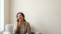 Woman with dark wavy hair, South Asian ethnicity, wearing patterned blouse, sitting in white chair, talking on phone, smiling, minimal interior setting.