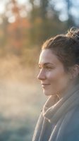 Young woman with messy bun hairstyle in cozy sweater enjoying peaceful moment outdoors with warm golden sunlight and soft bokeh background.