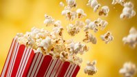 Fresh popcorn kernels bursting from red striped container against warm yellow background for movie theater snacks and entertainment concepts.