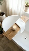 Wooden bath caddy with white waffle towel and lit candle draped over white freestanding tub, surrounded by potted plants and sheer curtains for spa relaxation.