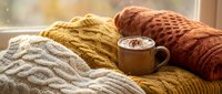 Cozy autumn scene with stacked knit sweaters in cream, mustard, and rust tones beside a warm beverage mug by a sunlit window, perfect for fall lifestyle and seasonal content.