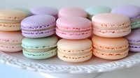 Pastel French macarons in delicate pink, mint, lavender and peach colors arranged on white decorative plate, showing sweet meringue cookies with filling.