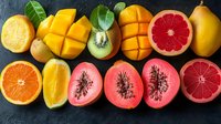 Fresh tropical fruits cut in halves showing vibrant colors and textures, including mango, citrus, papaya, kiwi arranged in rows on dark background. Perfect for healthy eating concept.
