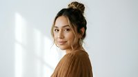 Young Hispanic woman with messy bun hairstyle wearing brown ribbed sweater looking over shoulder with natural makeup and soft lighting for lifestyle content.
