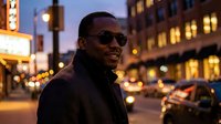 Man wearing sunglasses standing on urban street at dusk with illuminated storefronts and city lights in background, suitable for lifestyle and fashion content.