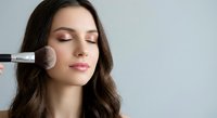 Young Caucasian woman applying blush with makeup brush, eyes closed, professional beauty routine for cosmetics and skincare advertising campaigns.