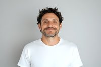 Middle-aged Mediterranean man with curly dark hair and stubble beard wearing white t-shirt smiling warmly at camera against light gray background. Genuine friendly expression.