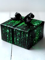 Gift box wrapped in black paper with glowing green matrix-style digital code pattern and elegant black ribbon bow on white wooden surface. Suitable for tech gifts.