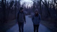 Two young adults walking on empty road through bare winter forest at dusk. Moody atmospheric scene with leafless trees and overcast sky for lifestyle content.