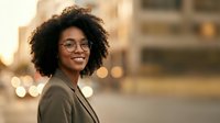 Young African American woman with curly hair and glasses wearing olive jacket, smiling outdoors at golden hour with blurred urban background, suitable for lifestyle and professional portraits.