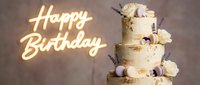 Three-tier birthday cake with gold leaf, macarons, white roses, and lavender, beside a glowing neon Happy Birthday sign on beige background.