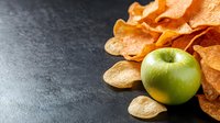 Fresh green apple with crispy potato chips scattered on dark stone background, healthy versus unhealthy food choice concept for snacking.