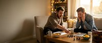 Two young men playing chess in cozy living room with warm lighting and candles creating intimate atmosphere for strategic board game competition.