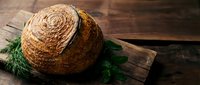 Freshly baked artisan sourdough bread loaf with golden crust on rustic wooden cutting board with fresh dill herbs for bakery and cooking concepts.