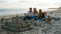 Caucasian family of four enjoying beach time together, sitting near sandcastle on sandy shore with ocean waves and coastal cliffs in background during golden hour.