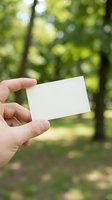 Hand holding a blank white card outdoors with green bokeh forest background, mockup for business card or invitation design.