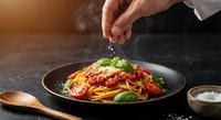 Chef hand sprinkling cheese over spaghetti pasta with tomato sauce and fresh basil in dark bowl on kitchen counter for gourmet Italian cuisine presentation.