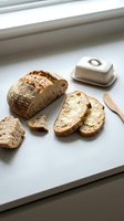 Freshly sliced artisan sourdough bread with crispy crust displayed on white surface near window with butter dish and wooden spreader for breakfast or bakery marketing.
