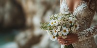 Bride in elegant lace dress holding delicate white daisy bouquet with greenery for romantic wedding ceremony celebration.