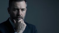 Thoughtful middle-aged Caucasian businessman in dark shirt with hand on chin in contemplative pose against gray background for corporate marketing.