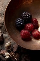 Fresh blackberries and raspberries in rustic copper bowl with dramatic lighting and dark moody background, styled with dried flowers for food photography.