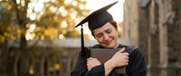 Young Caucasian woman in graduation cap and gown hugging diploma, crying with joy on university campus during commencement ceremony.