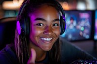 Young African American female gamer wearing purple headphones smiles while streaming or playing video games in dark room with neon lighting and computer setup.