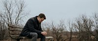 Young Caucasian man in dark jacket sitting alone on wooden bench in winter park, looking down pensively in contemplative mood during overcast day.