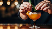 Bartender garnishing orange cocktail with fresh rosemary sprig in coupe glass at upscale bar with warm ambient lighting for mixology and hospitality.