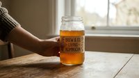 Caucasian woman's hand holding a glass mason jar with kombucha on a wooden table near a bright window.