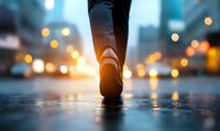 Walking shoe on wet pavement at dusk with glowing city lights creating atmospheric bokeh effect, low angle perspective captures reflection on rain-soaked street.