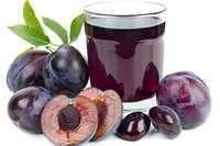 Plum juice in glass with fresh whole and halved plums with pit and green leaves on white background, suitable for beverage and food packaging.