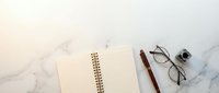 Minimalist workspace with open notebook, wooden pen, eyeglasses and small camera on white marble surface. Clean desk setup for planning, writing and creativity.