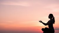 Woman practicing yoga meditation at sunset with peaceful pink sky creating serene wellness atmosphere for mindfulness and spiritual balance.