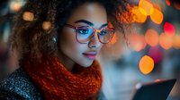 Young African American woman in glasses and orange scarf looking at digital tablet against blurred city lights background with bokeh effect, evening urban lifestyle.