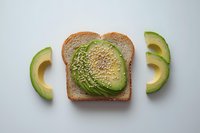 Sliced avocado on whole grain toast with sesame seeds, surrounded by curved avocado pieces on white background, fresh healthy breakfast composition.
