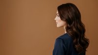 Young woman with long wavy brunette hair, fair complexion, wearing dark navy blue shirt, shown in profile against warm beige background. Suitable for beauty, haircare, and wellness marketing.