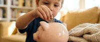 Young boy, approximately 5-8 years old, Caucasian ethnicity, placing coin into pink piggy bank. Indoor home setting with warm lighting, bookshelf and couch visible. Financial literacy and savings concept.