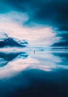 Lone figure stands on mirror-like glacial lake surface reflecting snow-capped mountains and dramatic turquoise sky, creating ethereal symmetrical landscape.