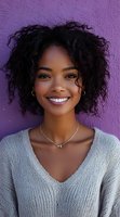 Young African American woman with natural curly hair smiling warmly against purple wall background. Wearing gray knit sweater, delicate jewelry, and radiating positive energy.