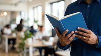 Man in navy shirt reading open blue book in modern office environment with blurred colleagues working at desks in background, suitable for education, learning, and professional development concepts.