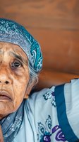 Elderly Southeast Asian woman wearing traditional blue patterned headscarf and white garment against warm terracotta background. Portrait captures weathered facial features and cultural attire for diversity and heritage themes.