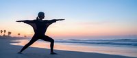 Woman practicing warrior yoga pose on beach at sunset with arms extended, silhouetted against colorful sky and ocean waves for wellness and meditation content.