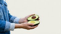 Young adult male with medium skin tone wearing denim shirt holding halved avocado with pit visible against white background. Suitable for healthy eating, nutrition, cooking, and food-related design projects.