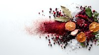 Aromatic spices and dried ingredients scattered on white surface with dramatic red powder trail, dried orange slices, bay leaves and berries creating culinary composition.