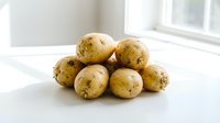 Fresh organic potatoes pile on white kitchen counter near window with natural daylight for healthy cooking and meal preparation at home.