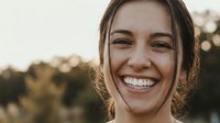 Young Caucasian woman with radiant smile and natural makeup enjoying golden hour sunlight outdoors with warm bokeh background for lifestyle content.