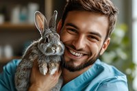 Young male veterinarian in blue scrubs holding grey rabbit, showing genuine smile while providing medical care. Close up portrait in clinic setting with natural lighting.